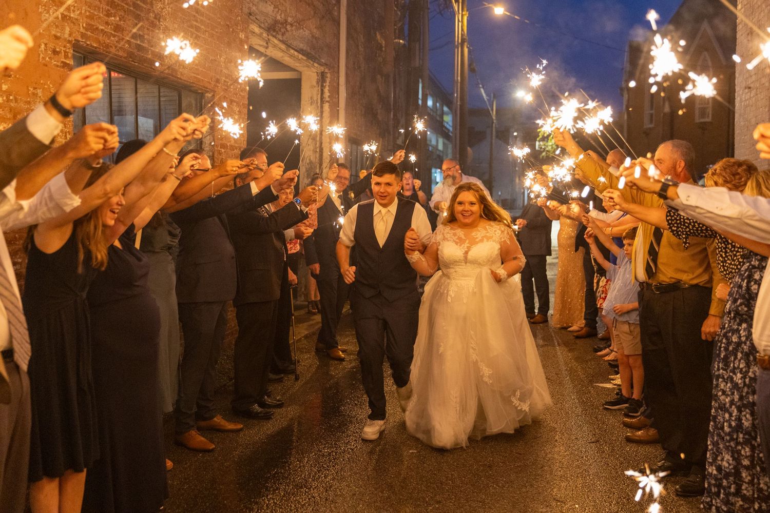Wedding guests line a street holding sparklers as the couple walks through the illuminated pathway.