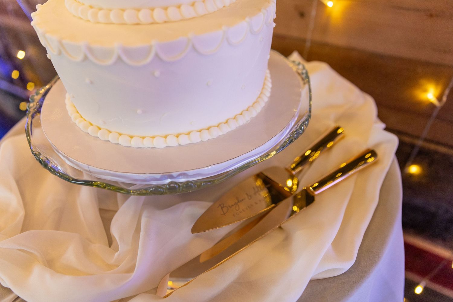 White tiered wedding cake with delicate piping on silver stand with gold serving utensils.