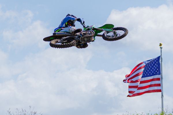 Motocross rider performs aerial jump with American flag waving in the background against blue sky.