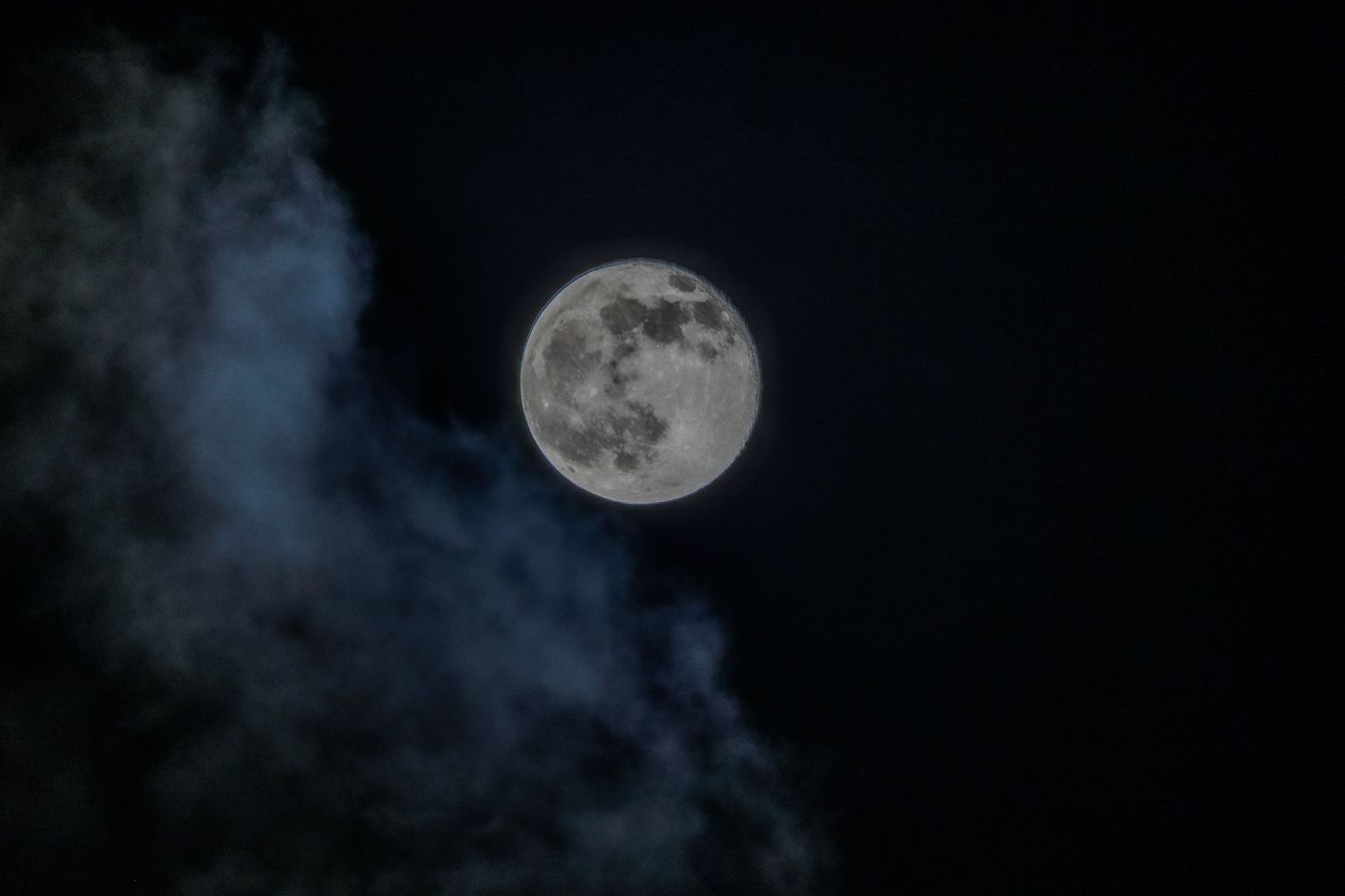 A bright full moon glows in a dark night sky surrounded by wispy blue-tinted clouds.