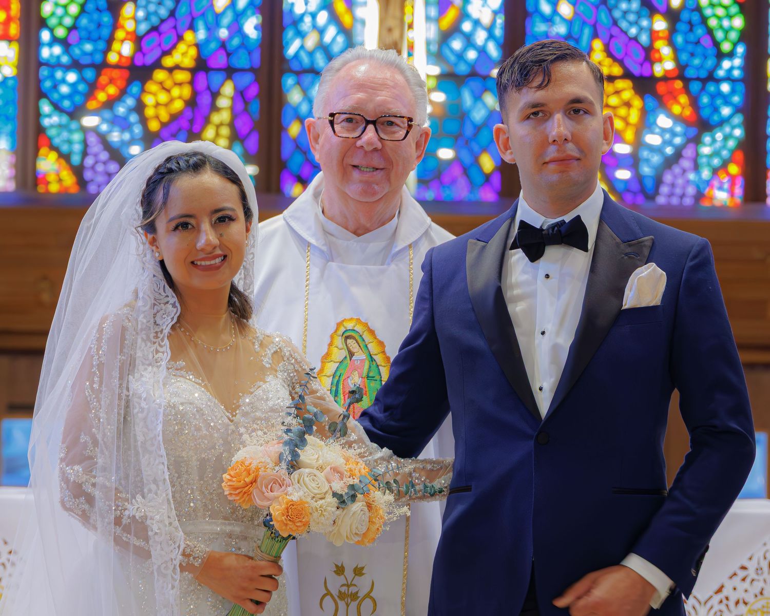 Wedding party poses in front of colorful stained glass windows in a church with peach and white floral bouquets.