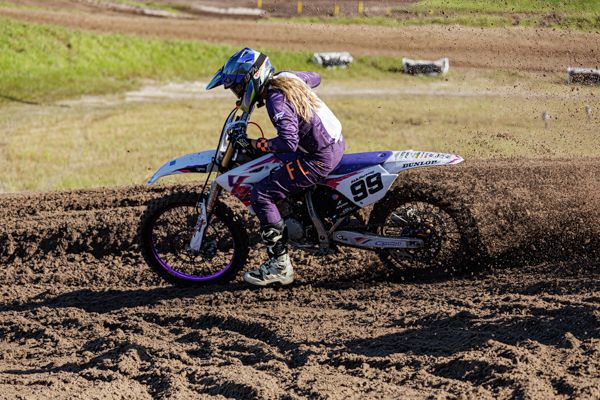 Dirt bike rider races through muddy motocross track kicking up soil during competitive race event.