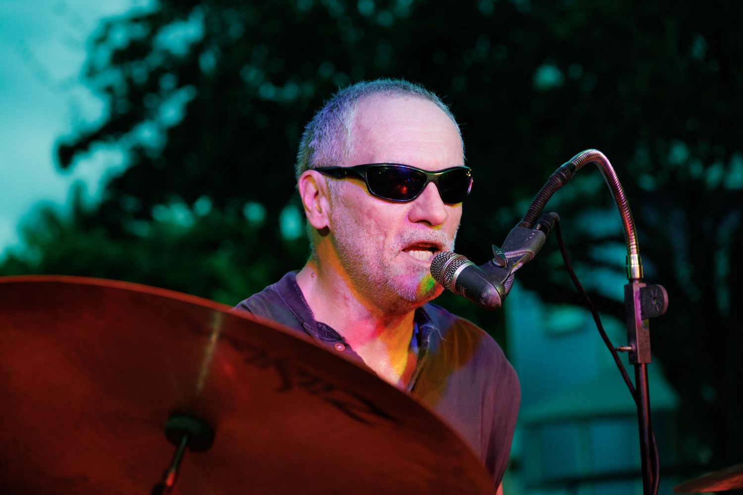 Musician in sunglasses performs on stage with a microphone during an outdoor concert.