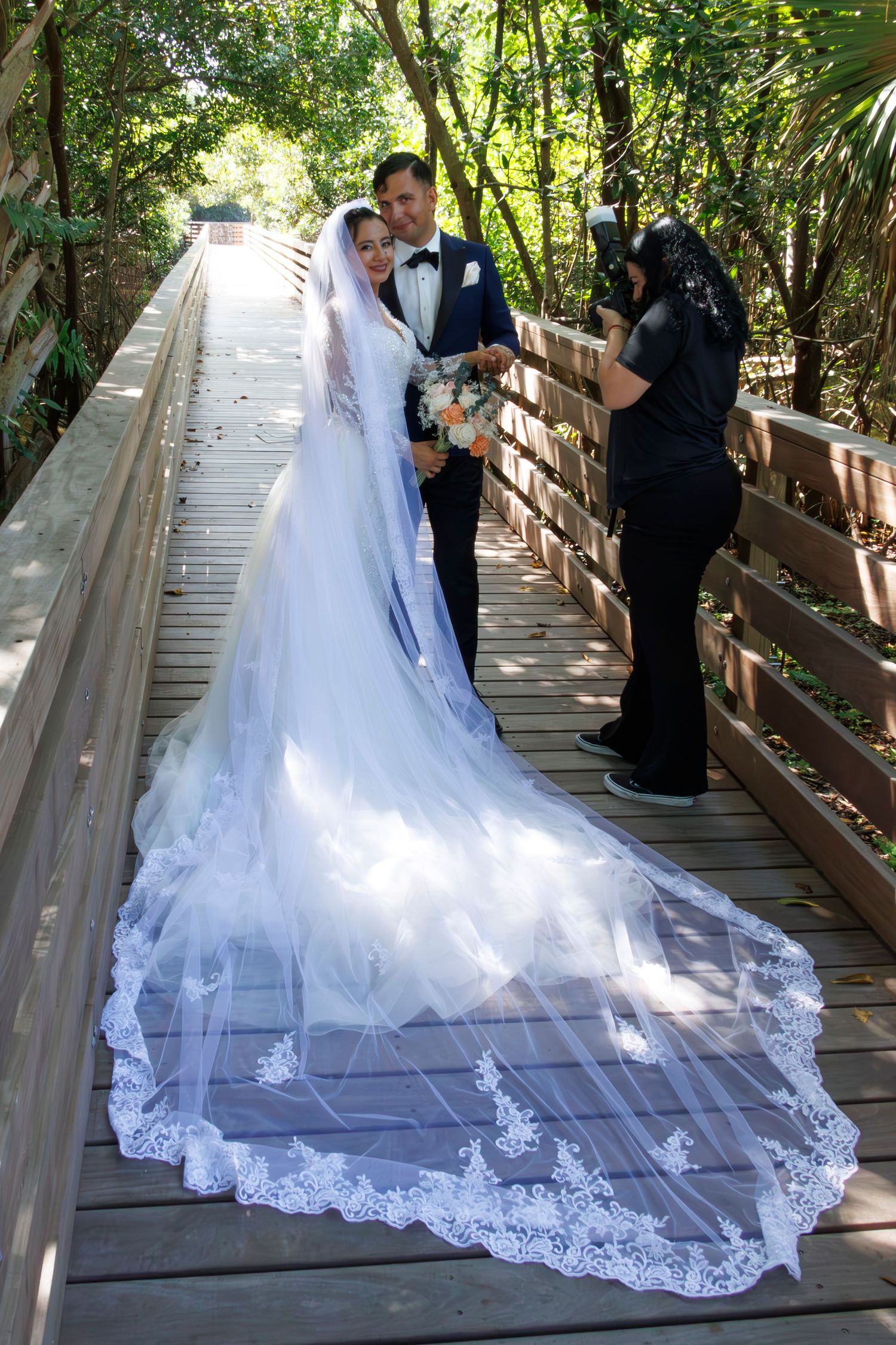 A couple in wedding attire stands on a wooden boardwalk surrounded by lush greenery as the bride's lace train trails behind.