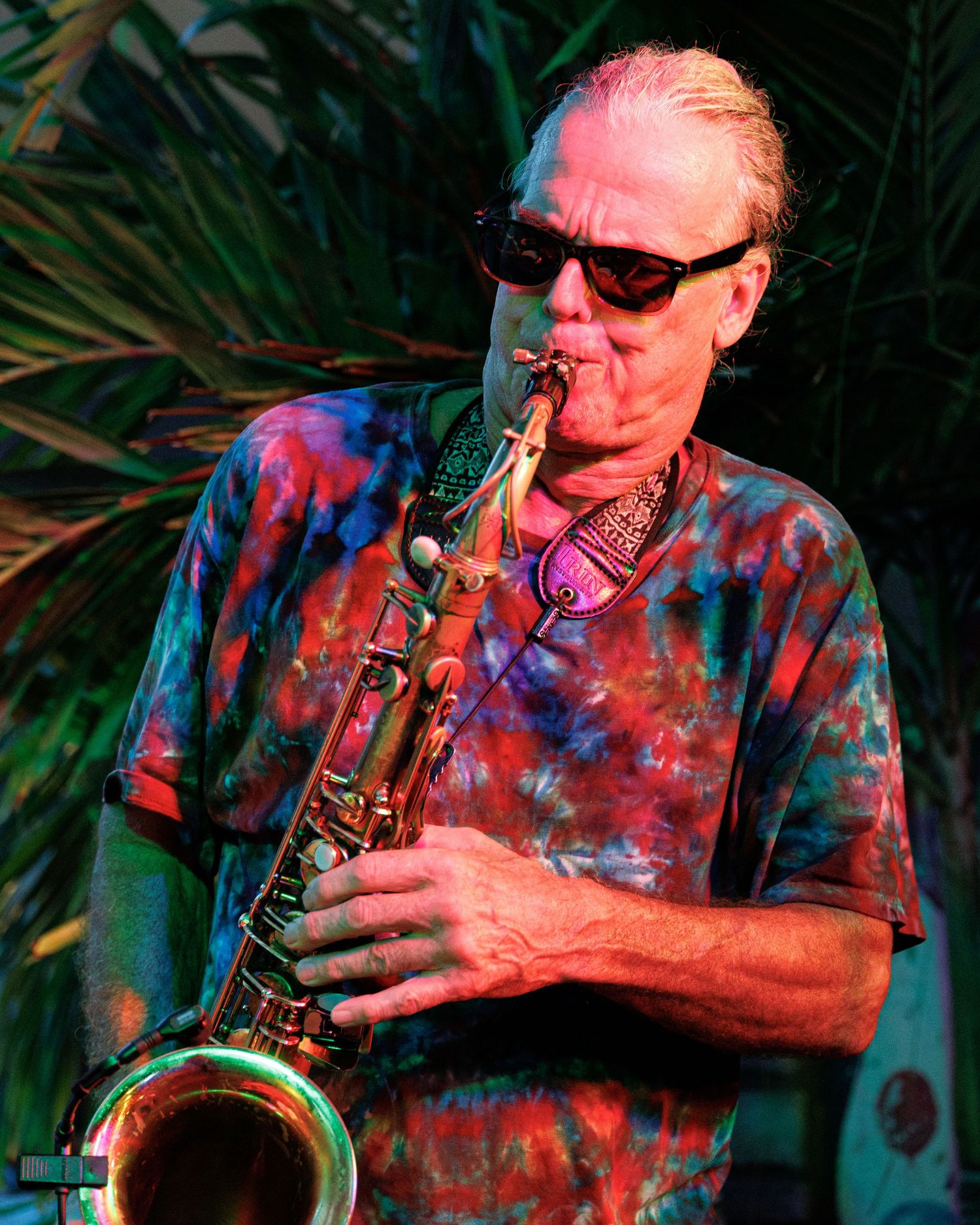 Jazz musician performs on saxophone wearing a colorful tie-dye shirt and sunglasses during an outdoor performance.