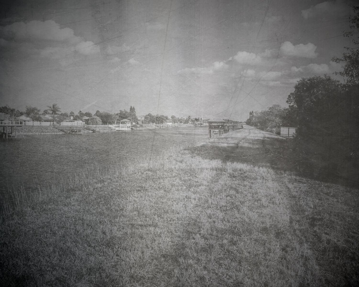 Vintage black and white photo of an open grassy field with small buildings and trees on the horizon under a cloudy sky.