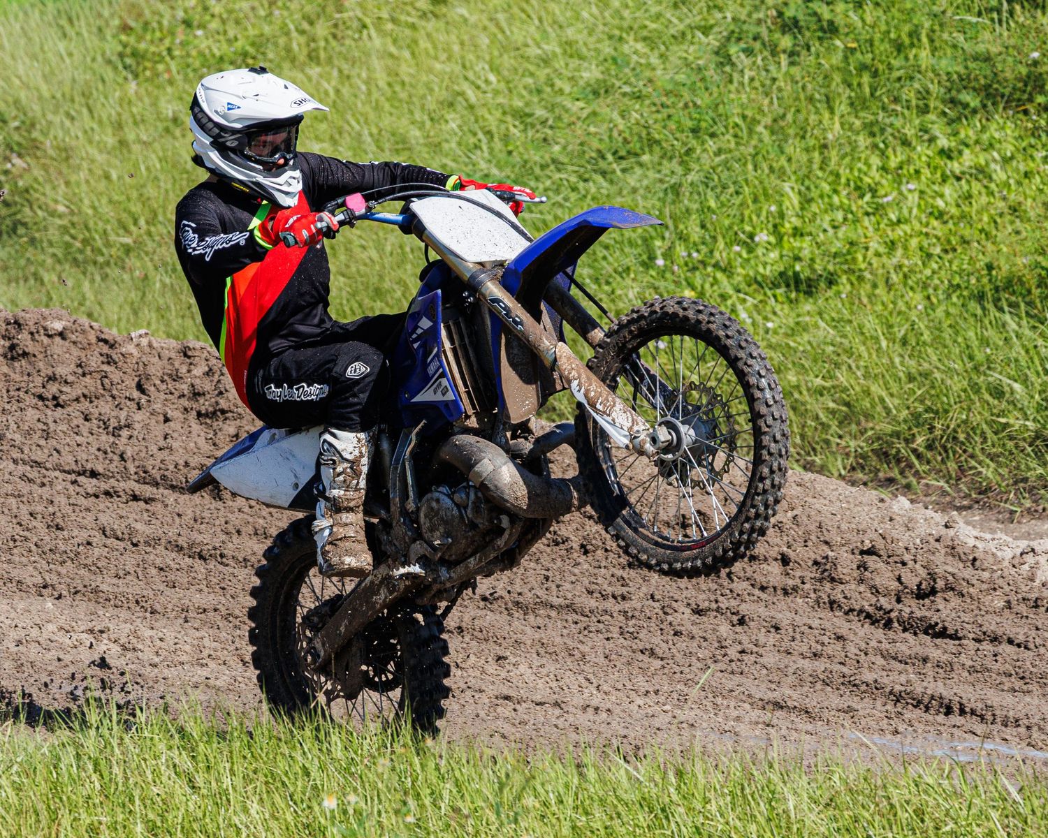 A dirt bike rider performs a wheelie on a muddy trail through green grass during off-road motorcycle riding.