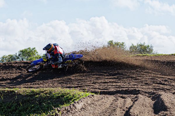 A motocross rider kicks up dirt while racing on a dirt track against a blue sky background.