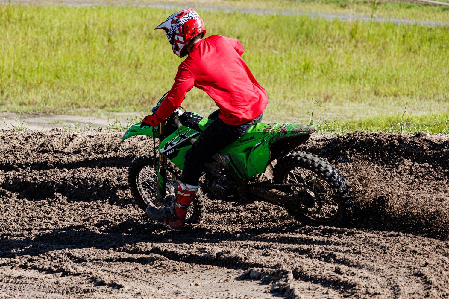 Motocross rider in red jacket and helmet navigates through muddy terrain on dirt bike.