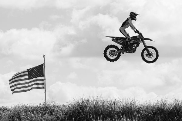 A silhouetted dirt bike rider jumps high against a cloudy sky with an American flag visible in the background.