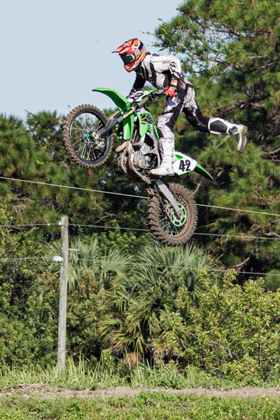Motocross rider performs impressive aerial jump on a green Kawasaki dirt bike against a backdrop of trees and bushes.