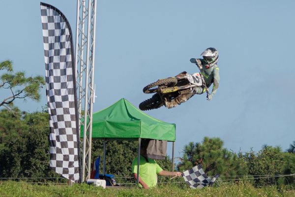 Motocross rider performs aerial backflip stunt on dirt bike against blue sky and checkered flag backdrop.
