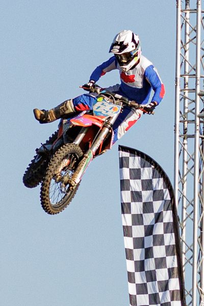Motocross rider performing aerial jump sequence against blue sky with checkered flag banners in background.