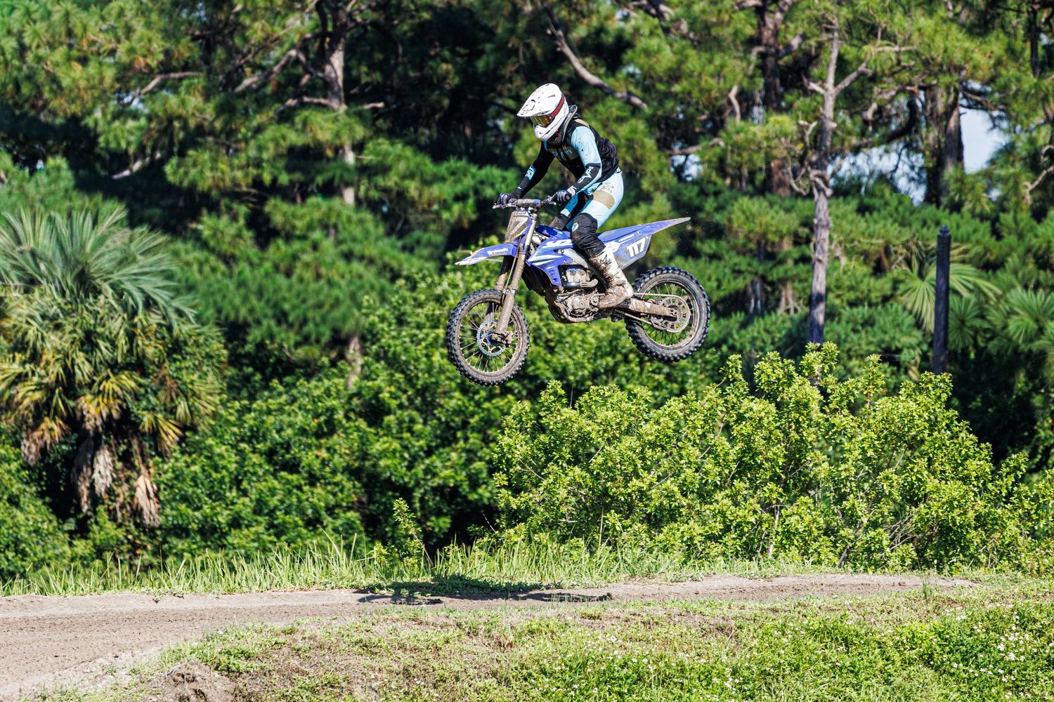 A dirt bike jumps high in the air over a grassy motocross track with lush green trees in the background.