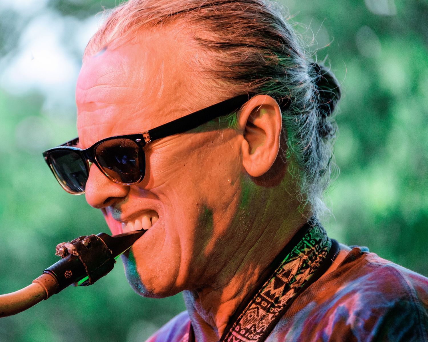 Close-up profile view of someone wearing sunglasses and smoking a pipe outdoors with green foliage in background.