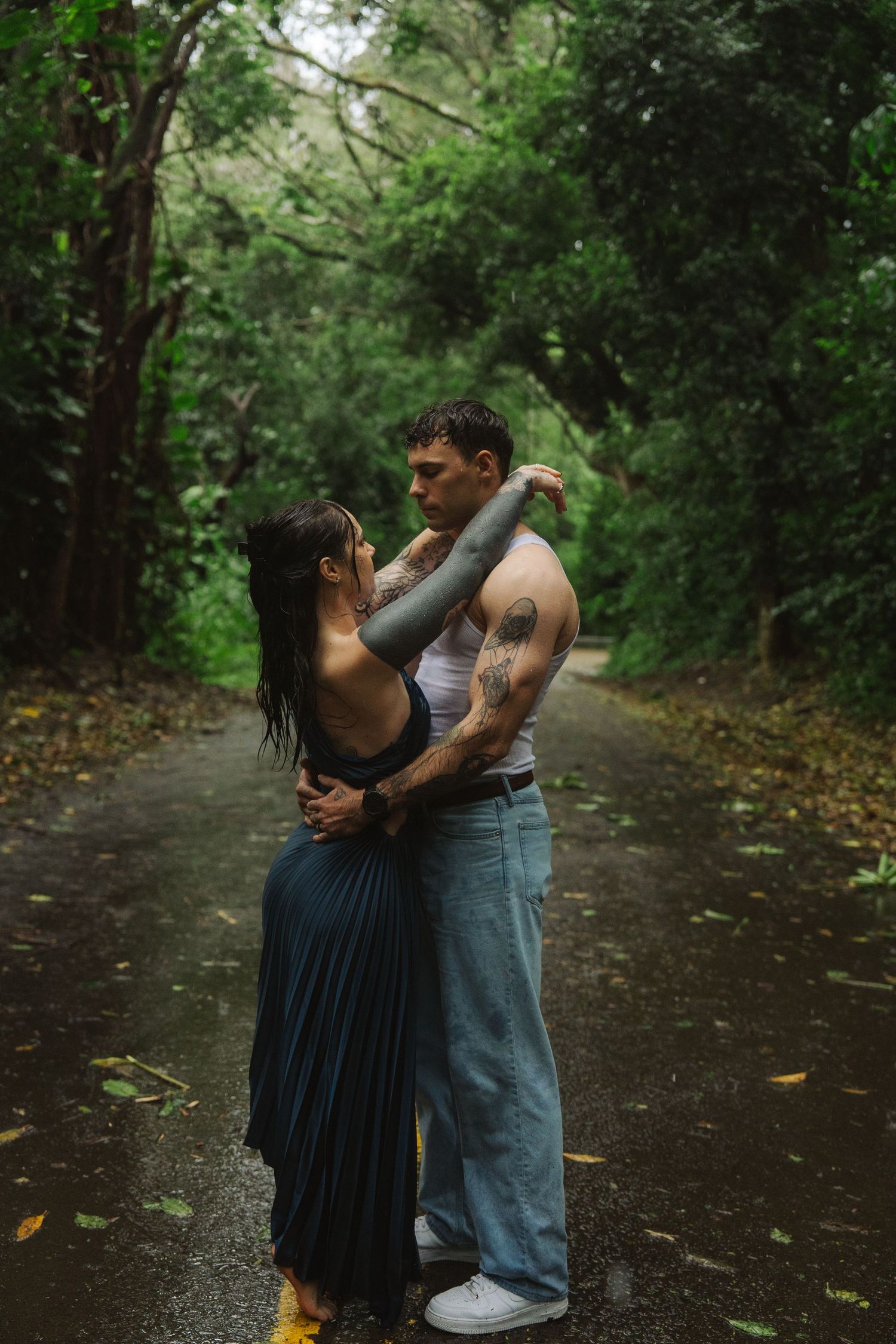 A tattooed couple embraces romantically on a rain-soaked forest road surrounded by lush green trees.