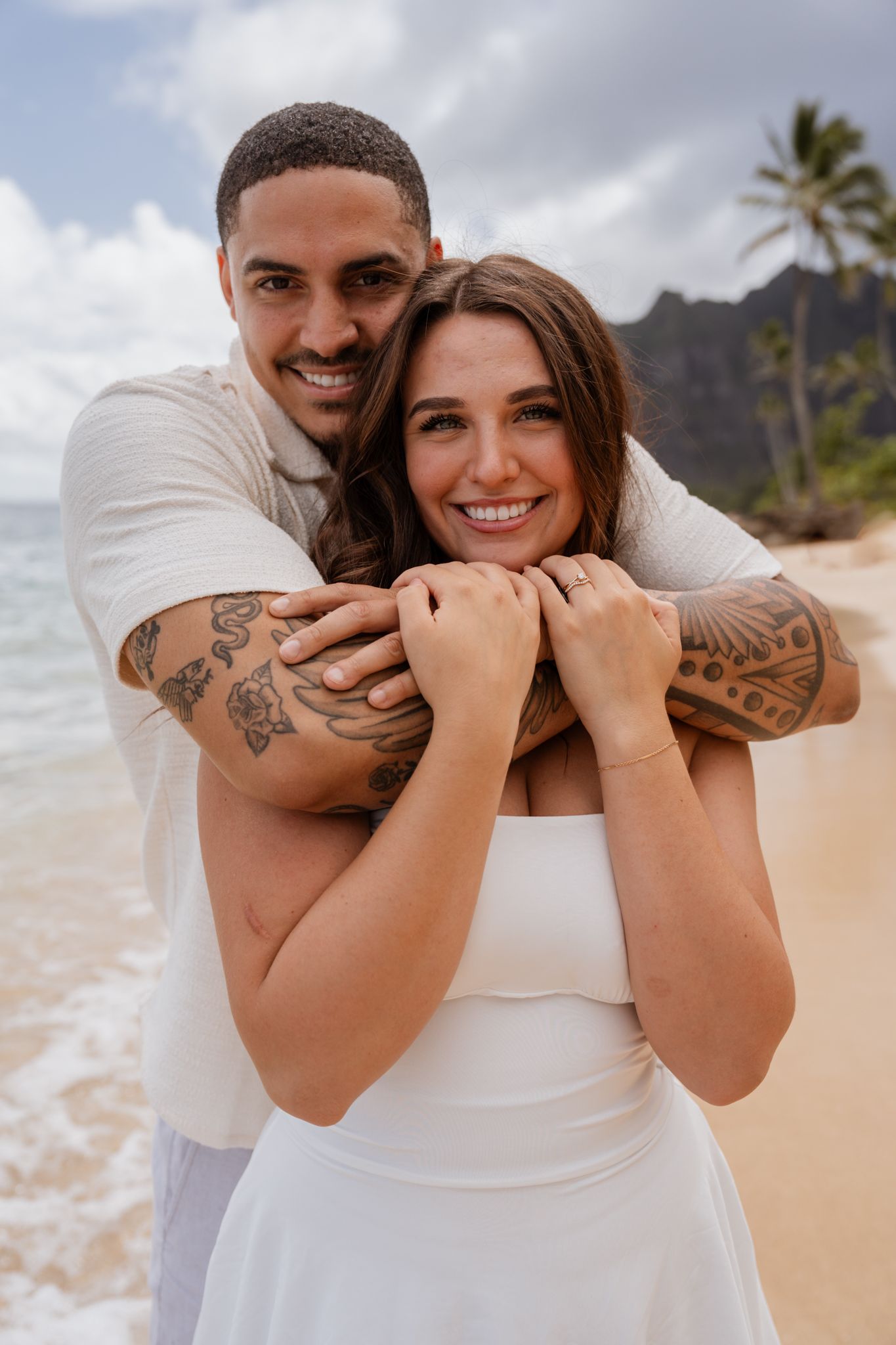 Joyful couple embracing on tropical beach with mountain backdrop.
