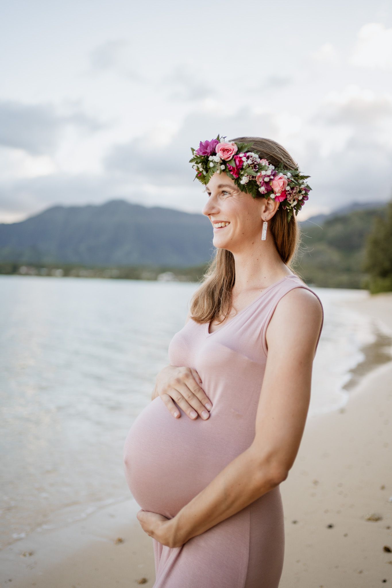 Expectant mother in pink dress wearing floral crown poses on tropical beach.