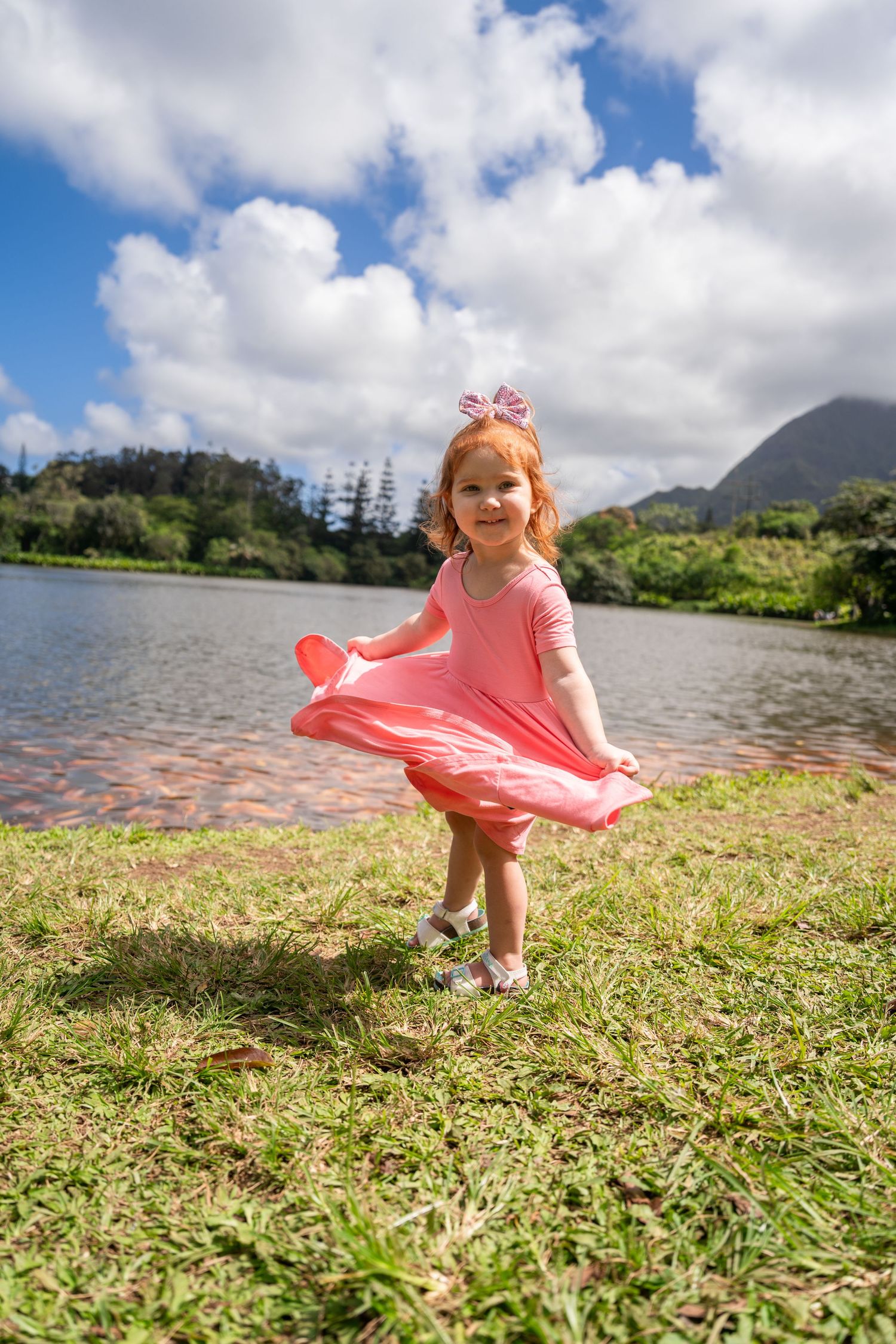 A young child in a pink dress twirls by a scenic mountain lake on a cloudy day.