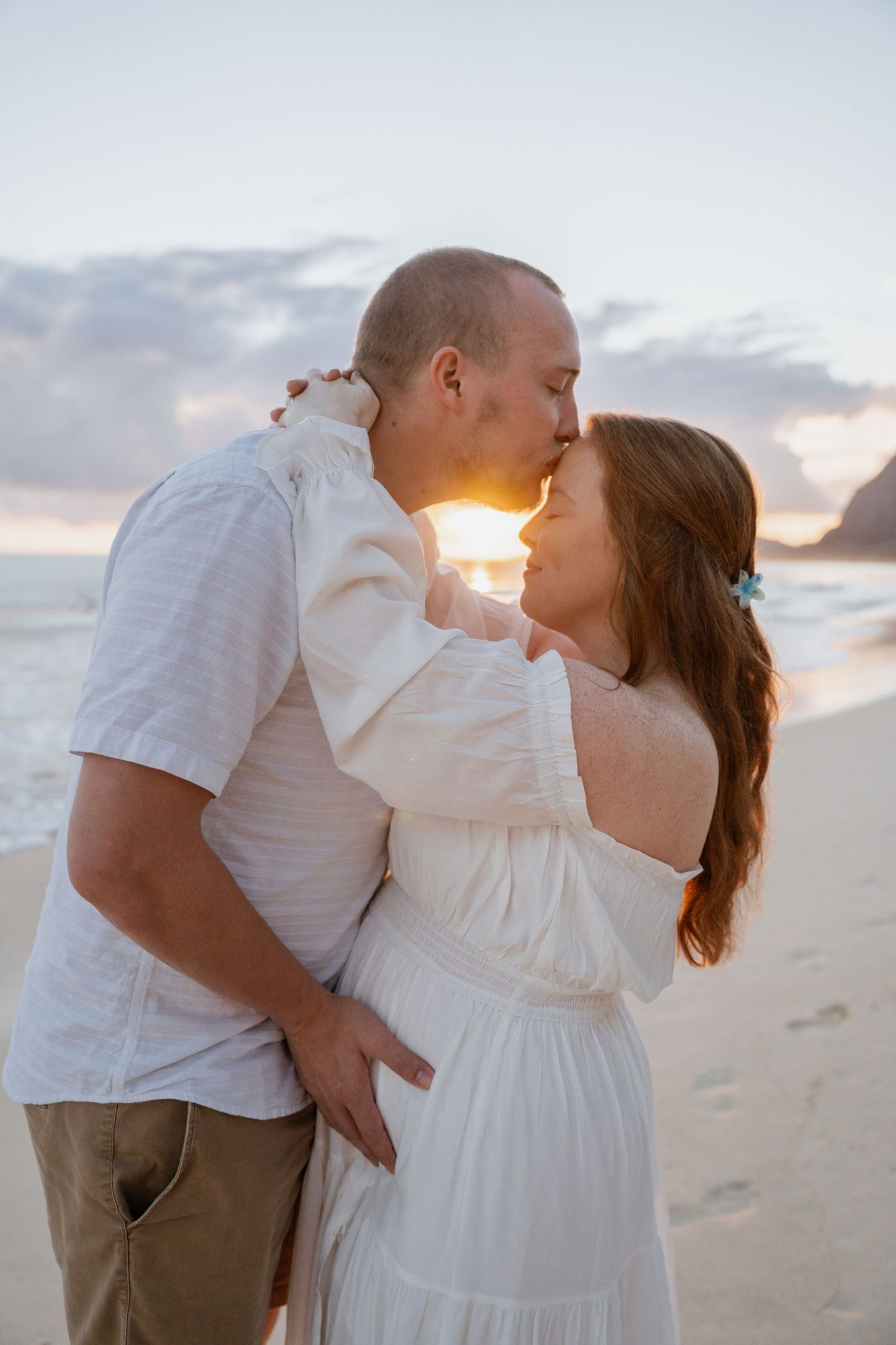 A romantic couple embraces and kisses on a sandy beach at sunset wearing white clothing.