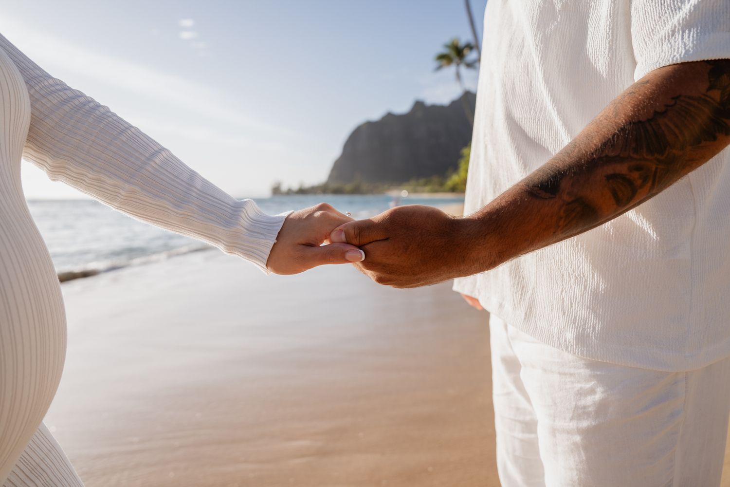 Close-up of two hands clasping together on a sunny beach with mountains in the background.