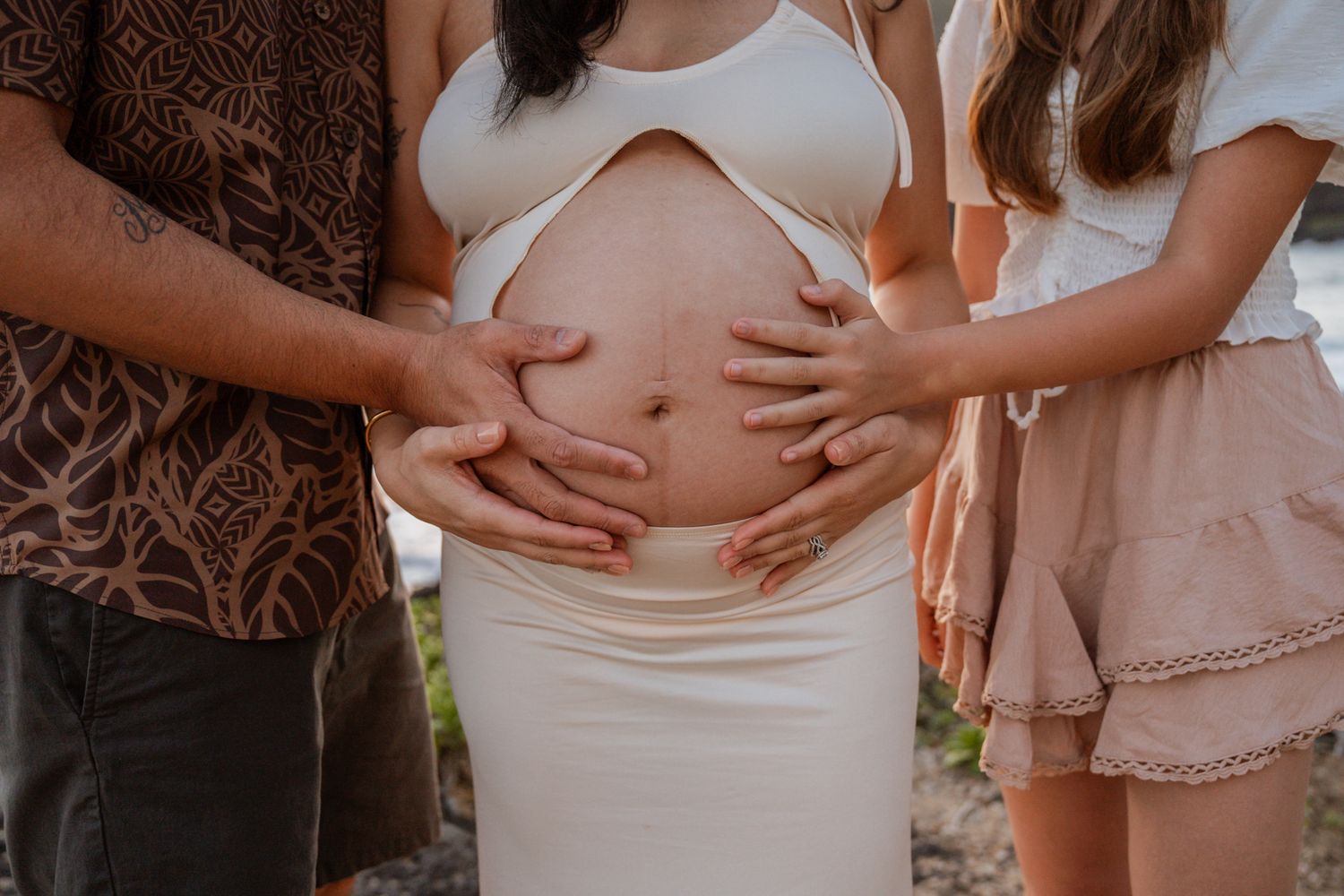 Multiple hands lovingly touching a pregnant belly in white dress on beach.