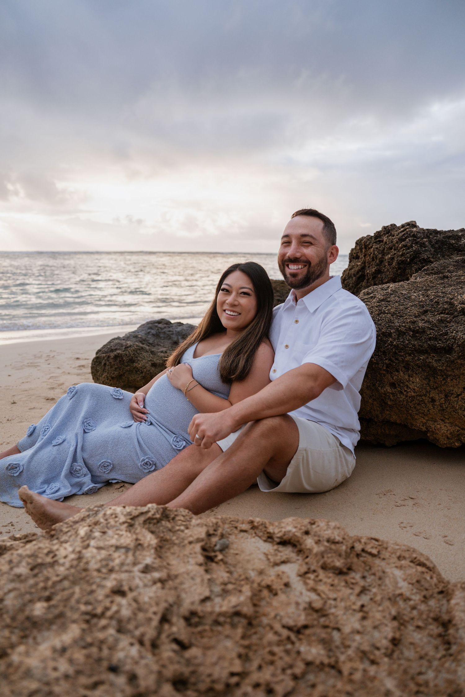 A couple in coordinated blue and white outfits share intimate moments while relaxing on a sandy beach at sunset.