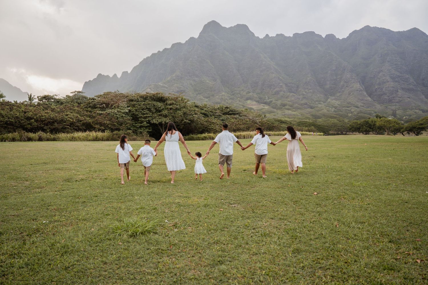 Group in white dresses holding hands and running across a grassy field with majestic mountains in the background in Hawaii.