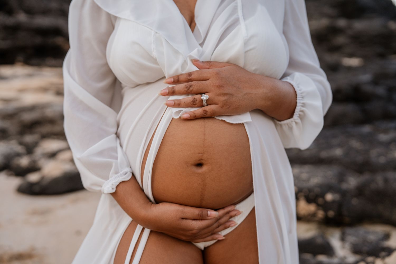 Close up of pregnant belly in white dress against rocky beach backdrop.