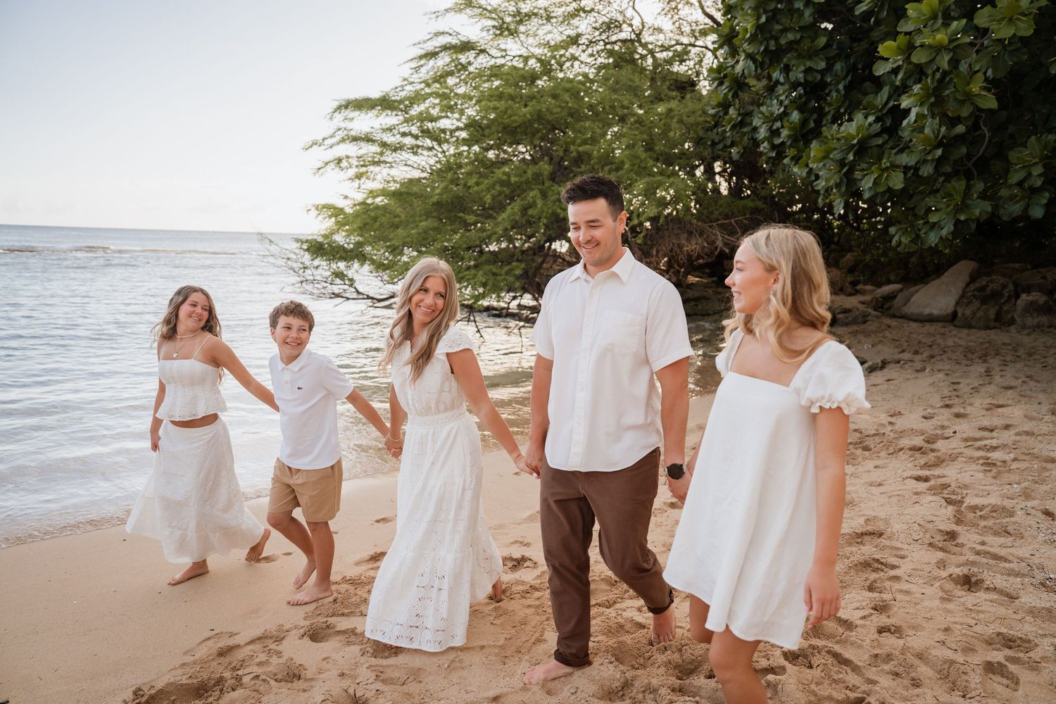 Family of five walking on sandy beach in white outfits, holding hands near ocean and tropical trees.