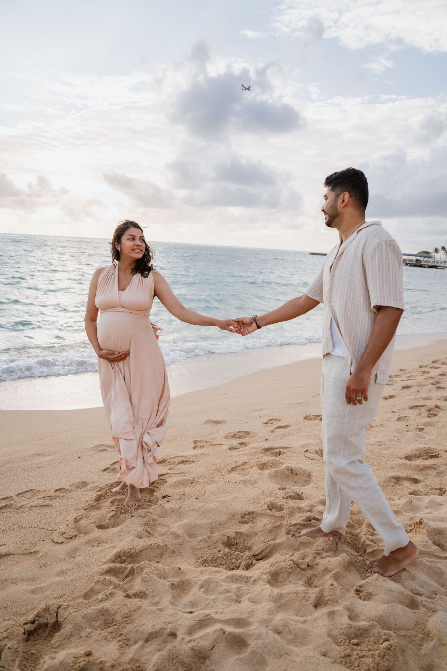 Pregnant couple holding hands on a sandy beach at sunset, woman in pink dress cradling baby bump.