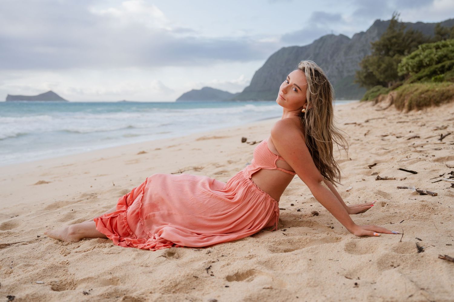 Woman in pink dress sitting on sandy beach with mountains and ocean in background.