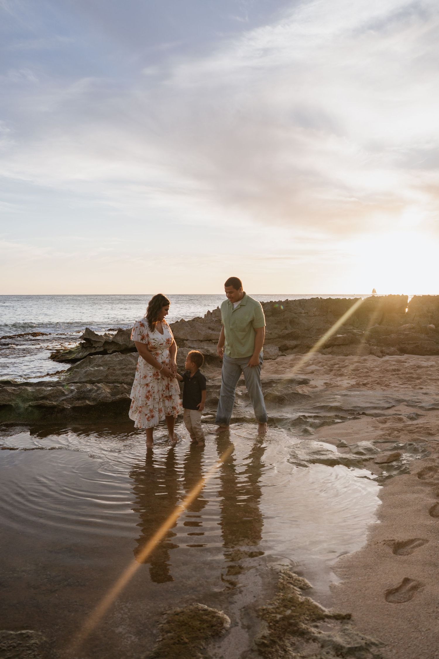 Family of three wading in shallow beach tide pools at golden hour sunset, parents holding toddler's hands near rocky shoreline.