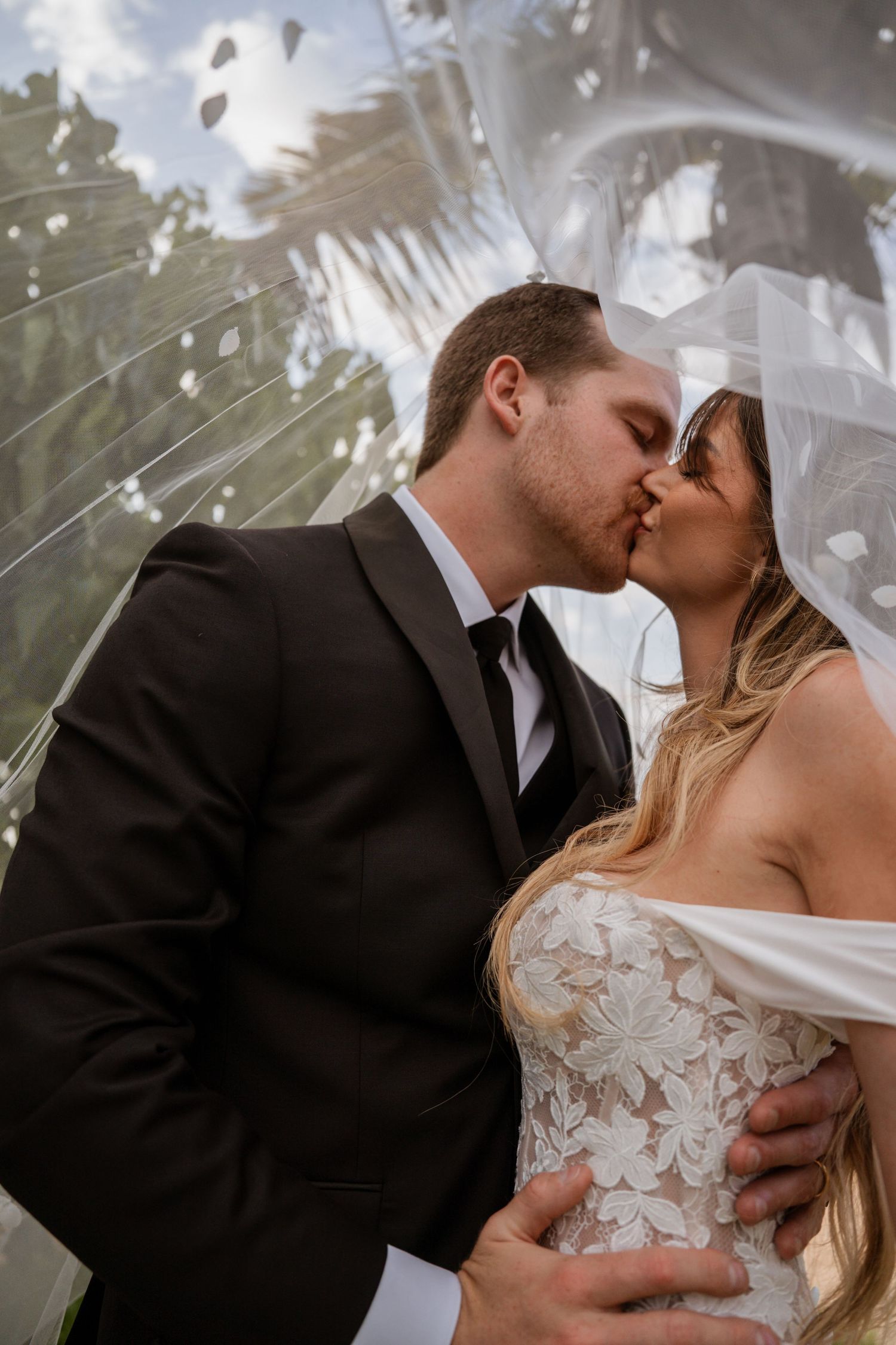 A bride and groom share a romantic kiss under a flowing veil at their outdoor wedding ceremony.
