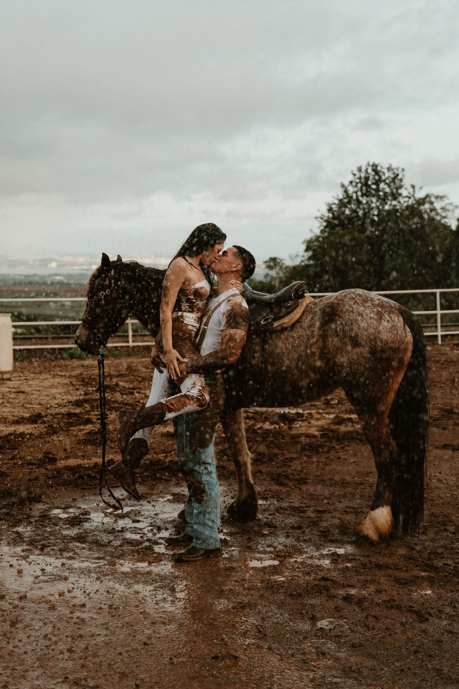 Couple kissing in the rain beside a horse in a muddy corral, romantic outdoor photoshoot.