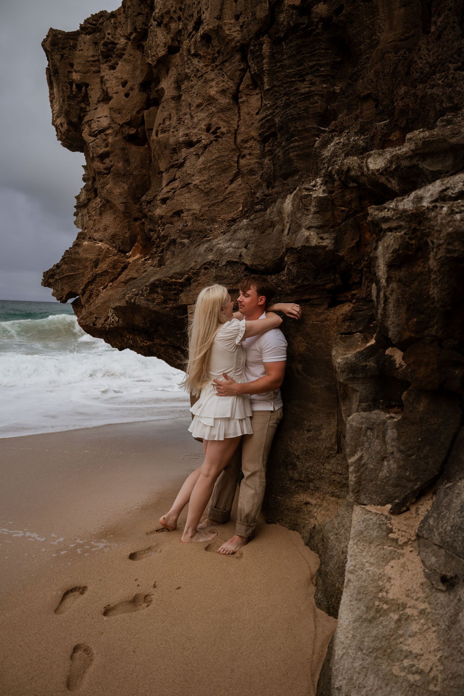 A couple embraces against a dramatic rocky cliff face on a beach with crashing waves and footprints in the sand.