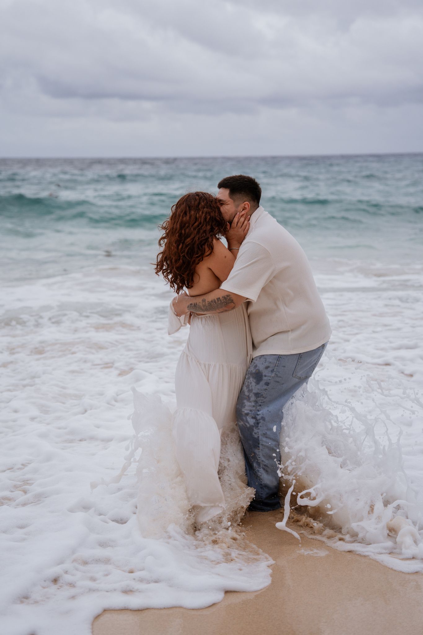 Romantic beach moment with waves crashing around couple in white attire.