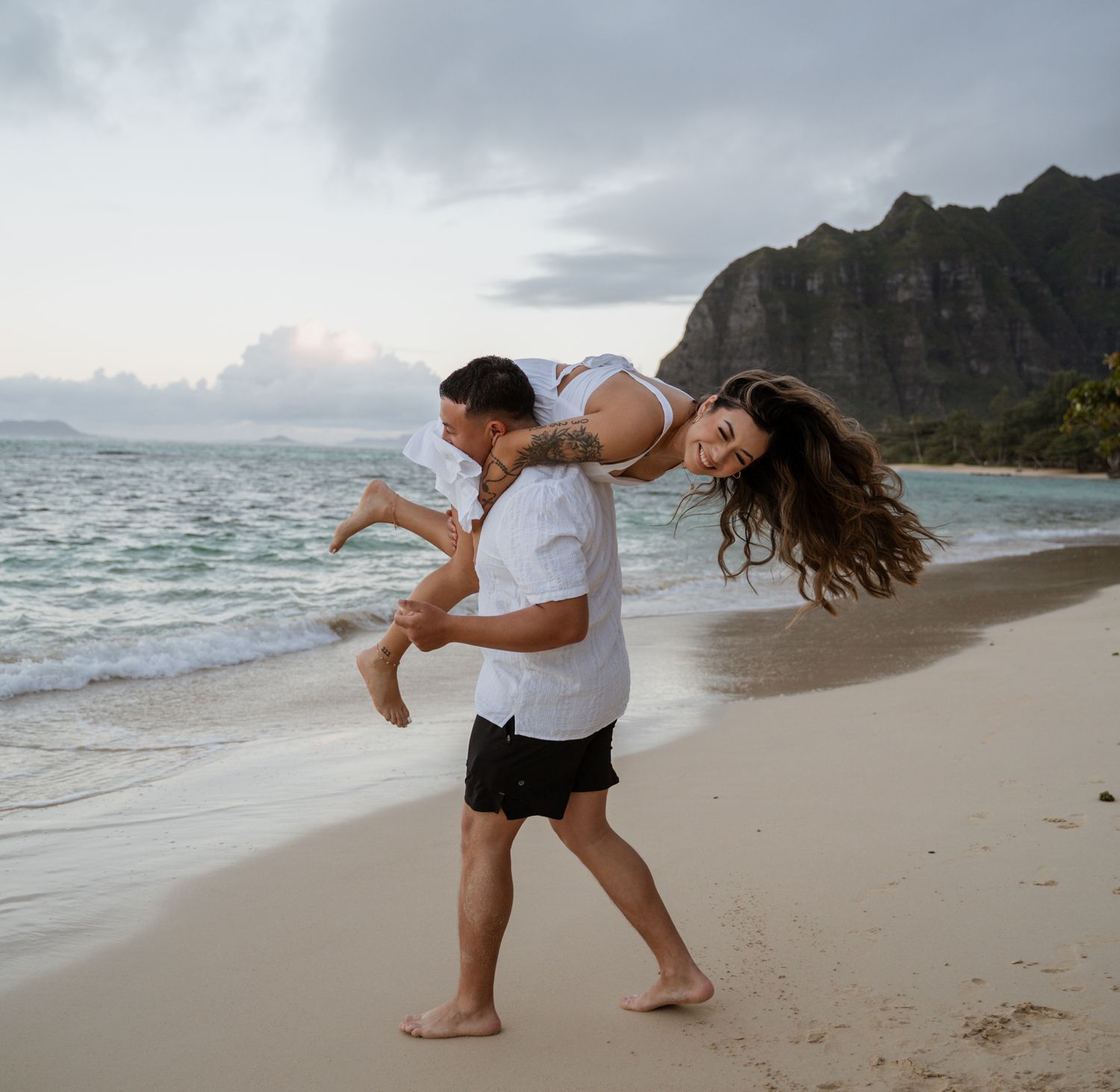 A playful couple enjoys a piggyback ride on a scenic Hawaiian beach with mountain views.