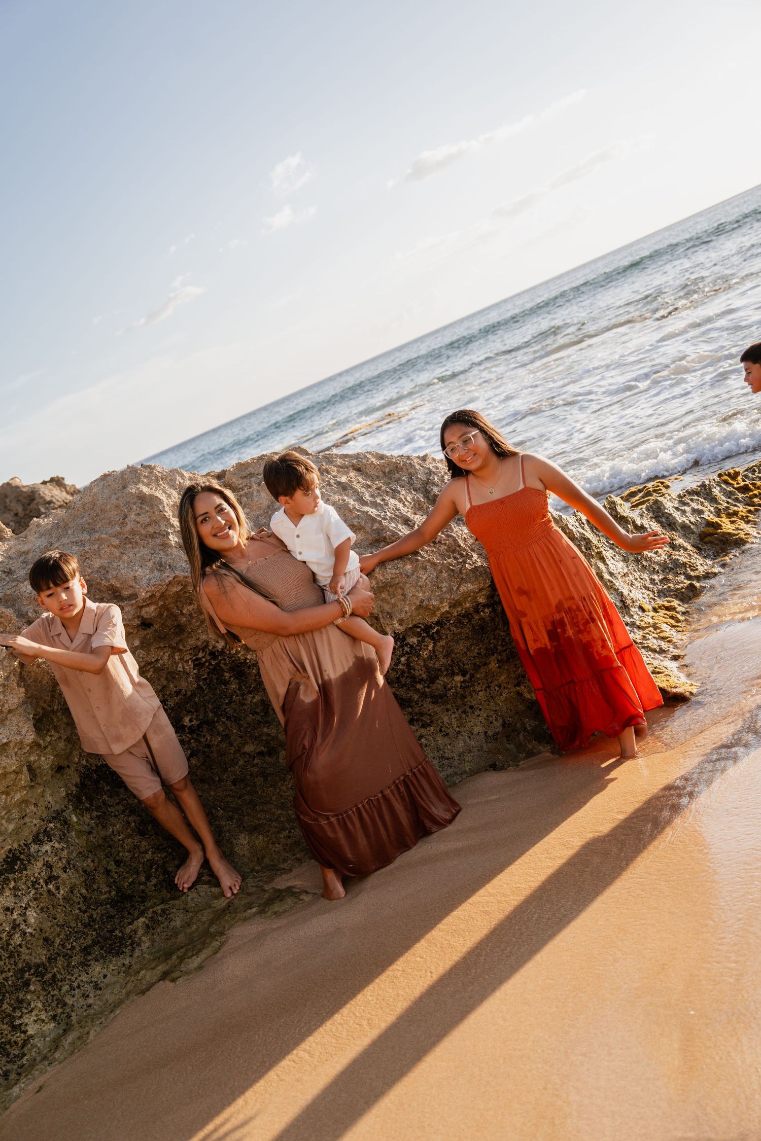 Family posing on sandy beach near rocks with ocean waves, wearing earth tones and red dress in golden sunlight.