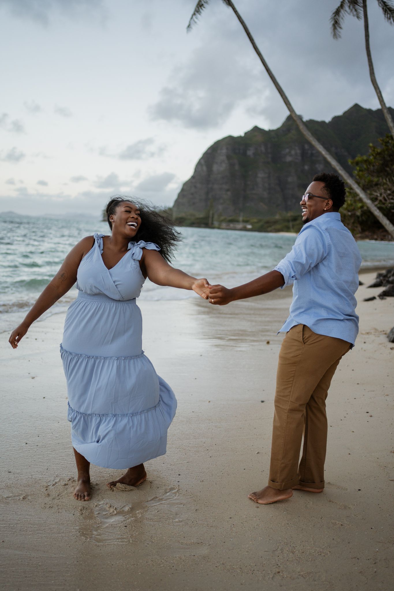 A couple dancing joyfully on a beach with dramatic mountain cliffs and palm trees in the background.