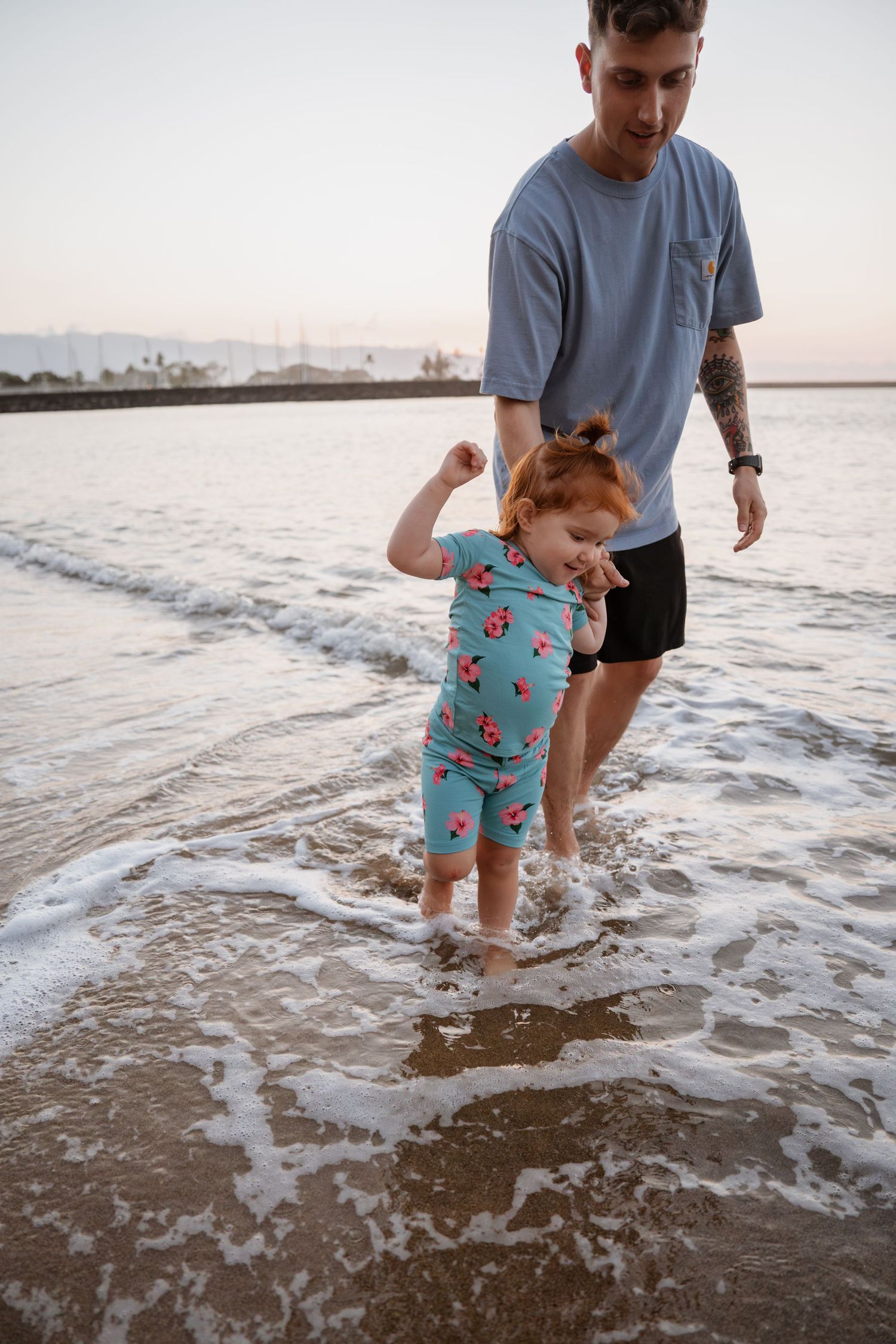 A parent and child splash in the ocean waves at sunset along a sandy beach.