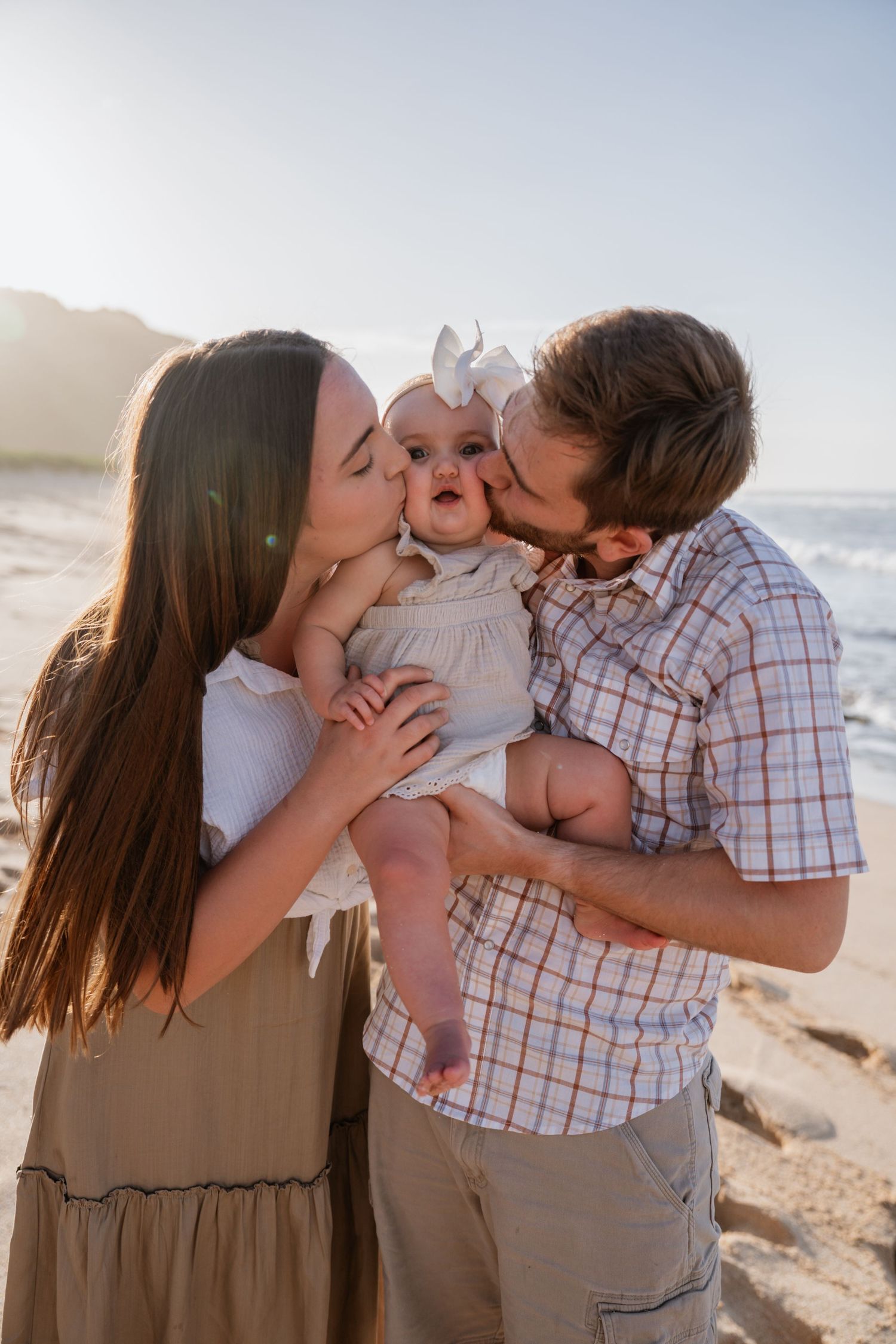 A couple kisses their baby girl on the cheeks at a sunny beach, creating a heartwarming family portrait.