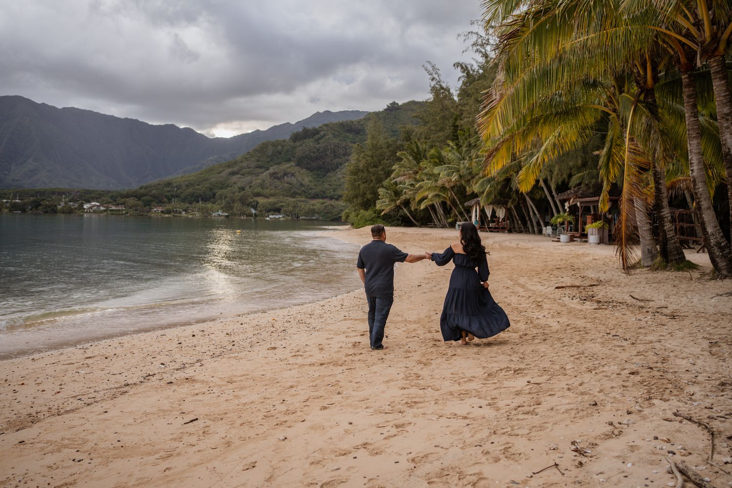 Two people walking hand in hand along a tropical beach with palm trees and mountains.