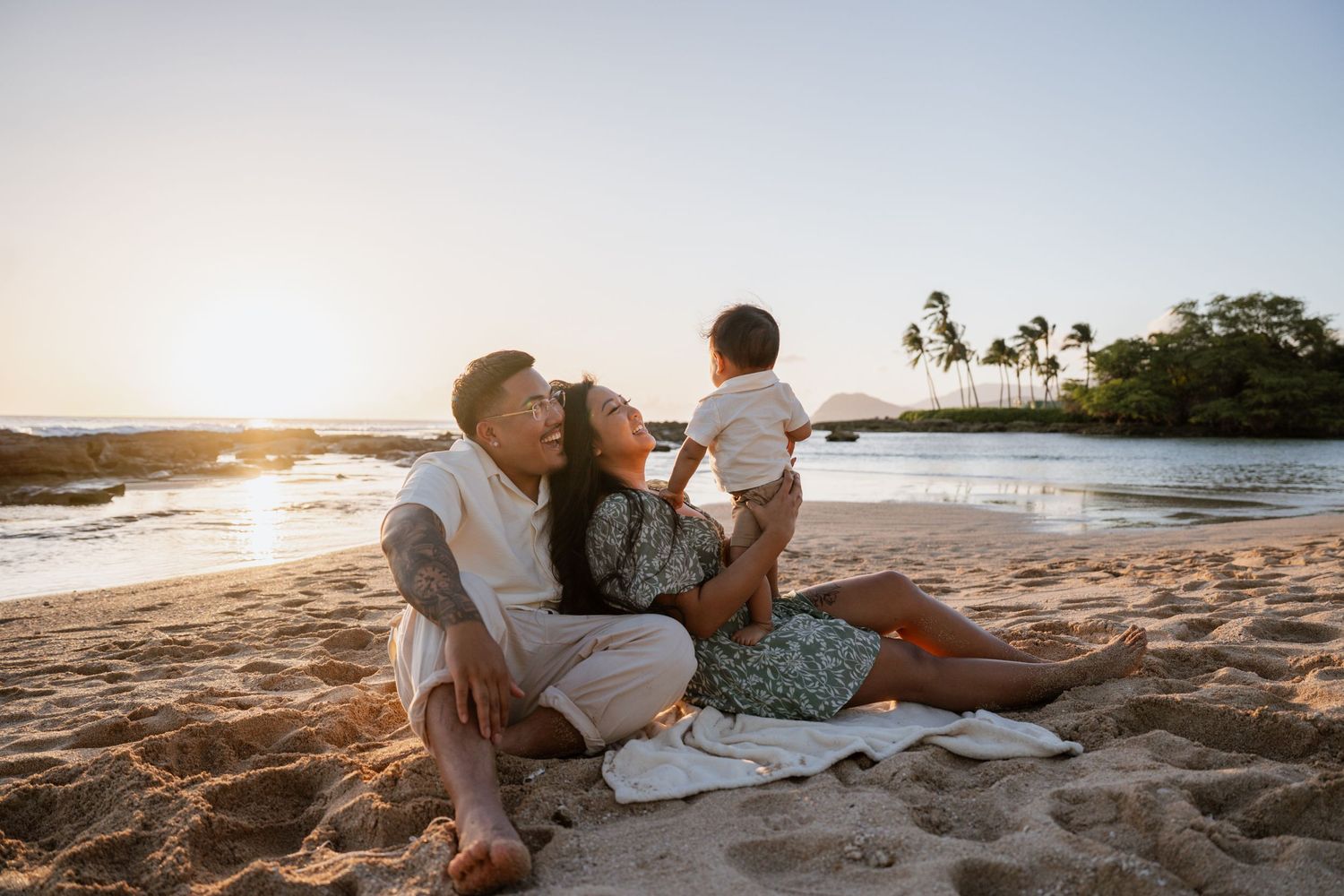 Family sitting on a sandy beach at sunset, parents lovingly gazing at their baby in a tropical setting.