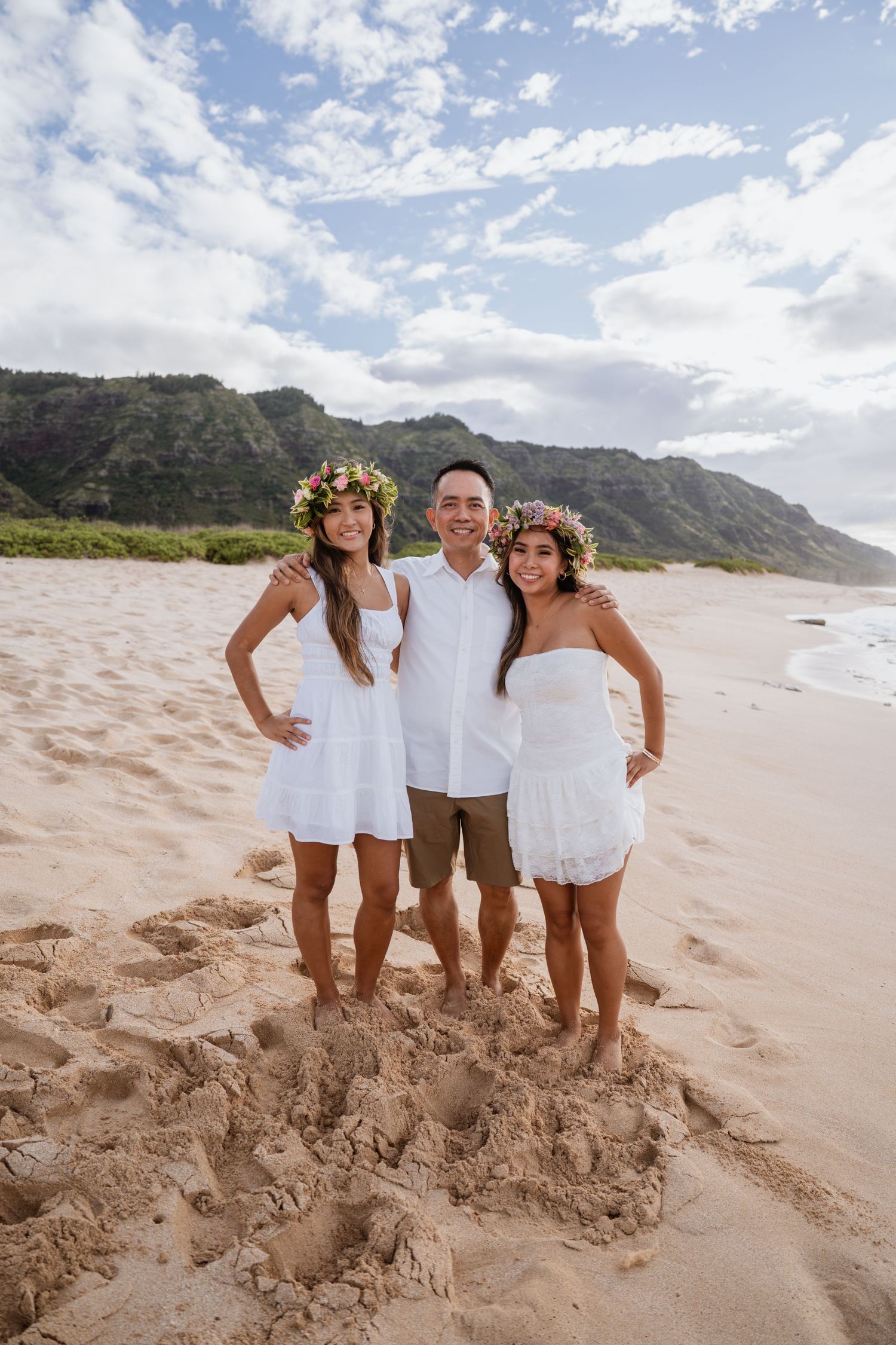 Group portrait on sandy Hawaiian beach with mountains and blue sky in the background showing matching white outfits.