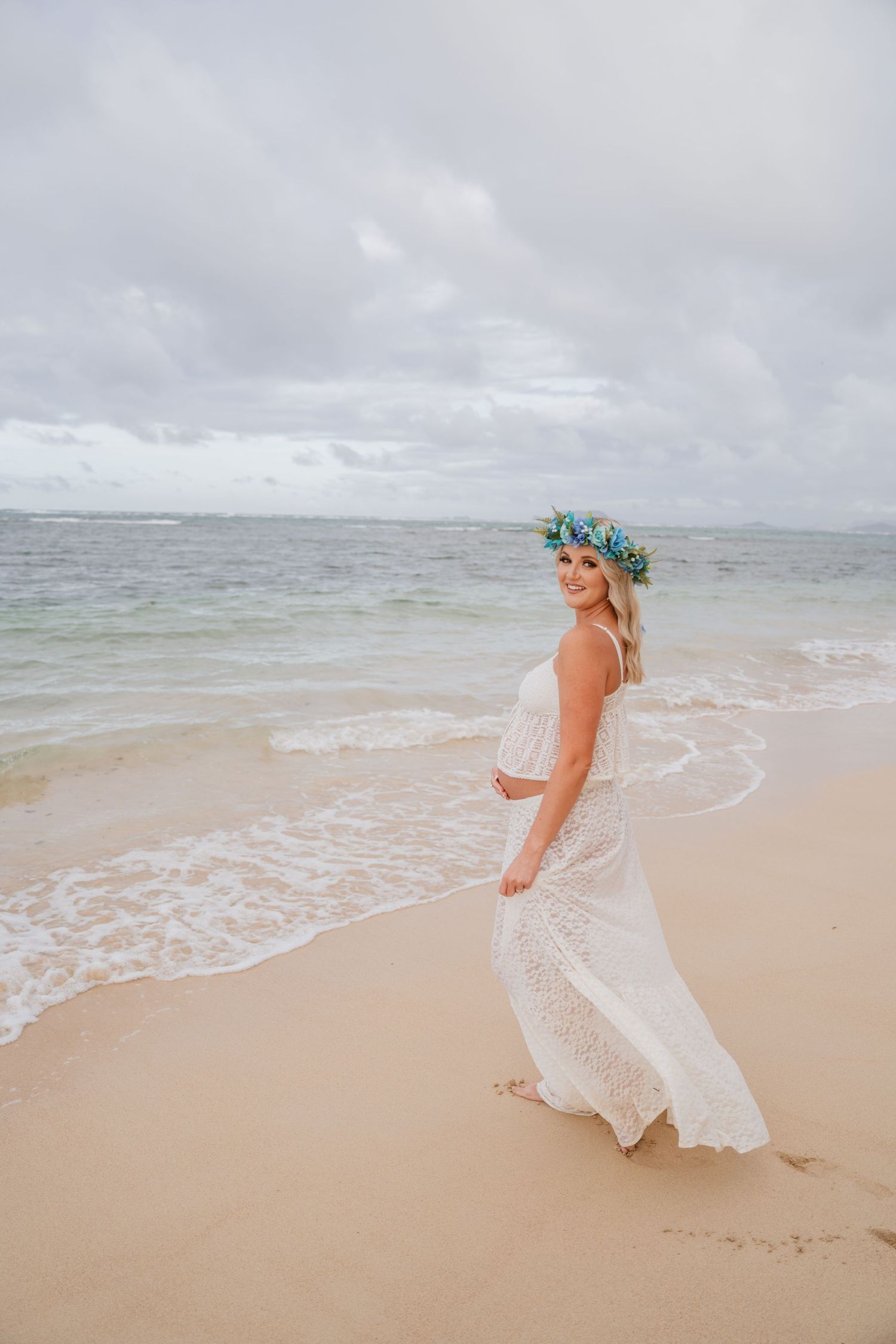Side view of person in flowing white dress and floral crown walking on beach with ocean waves and cloudy sky.