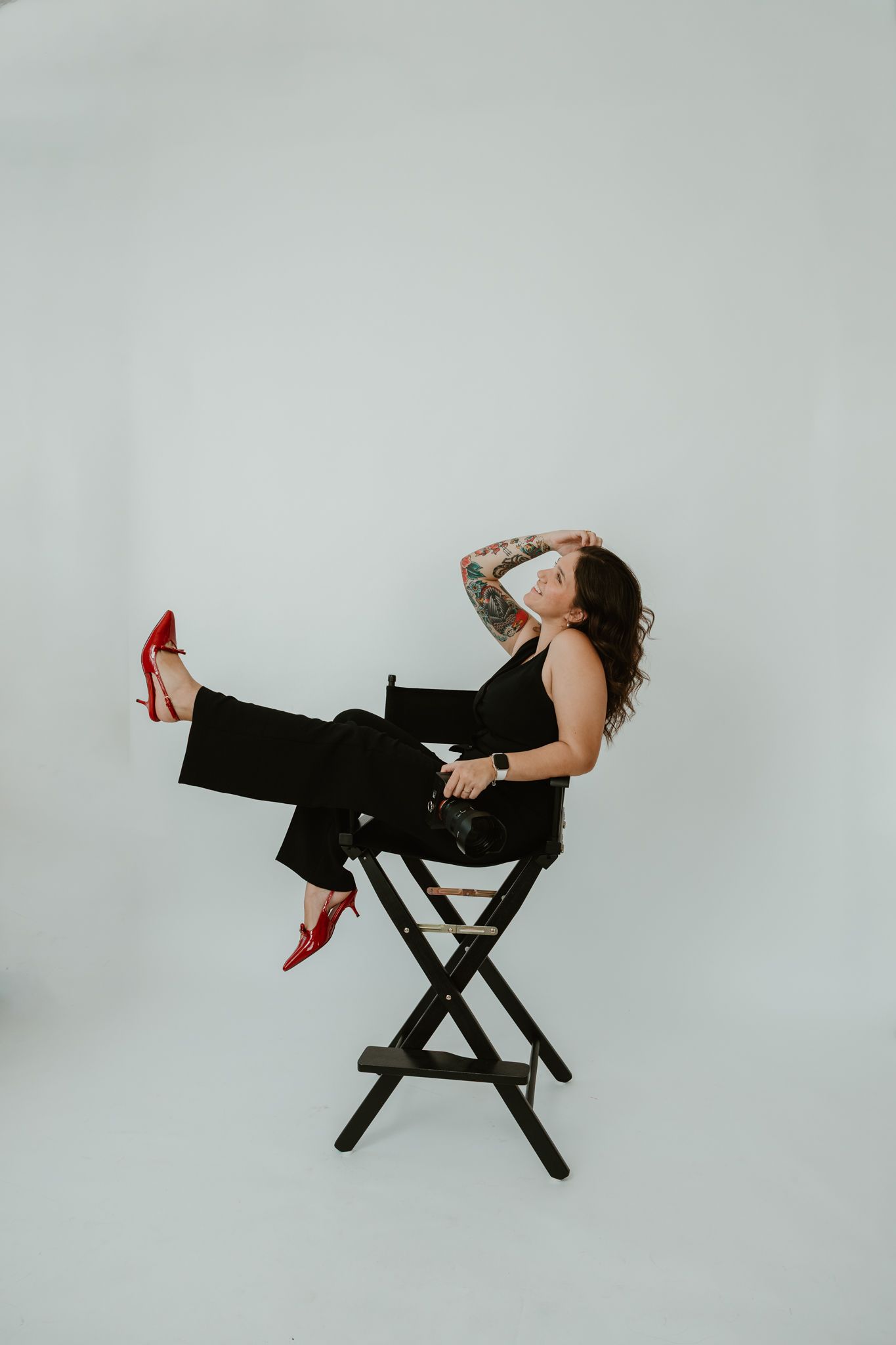 Stylish portrait of person in black outfit and red heels posing on director's chair against white background.