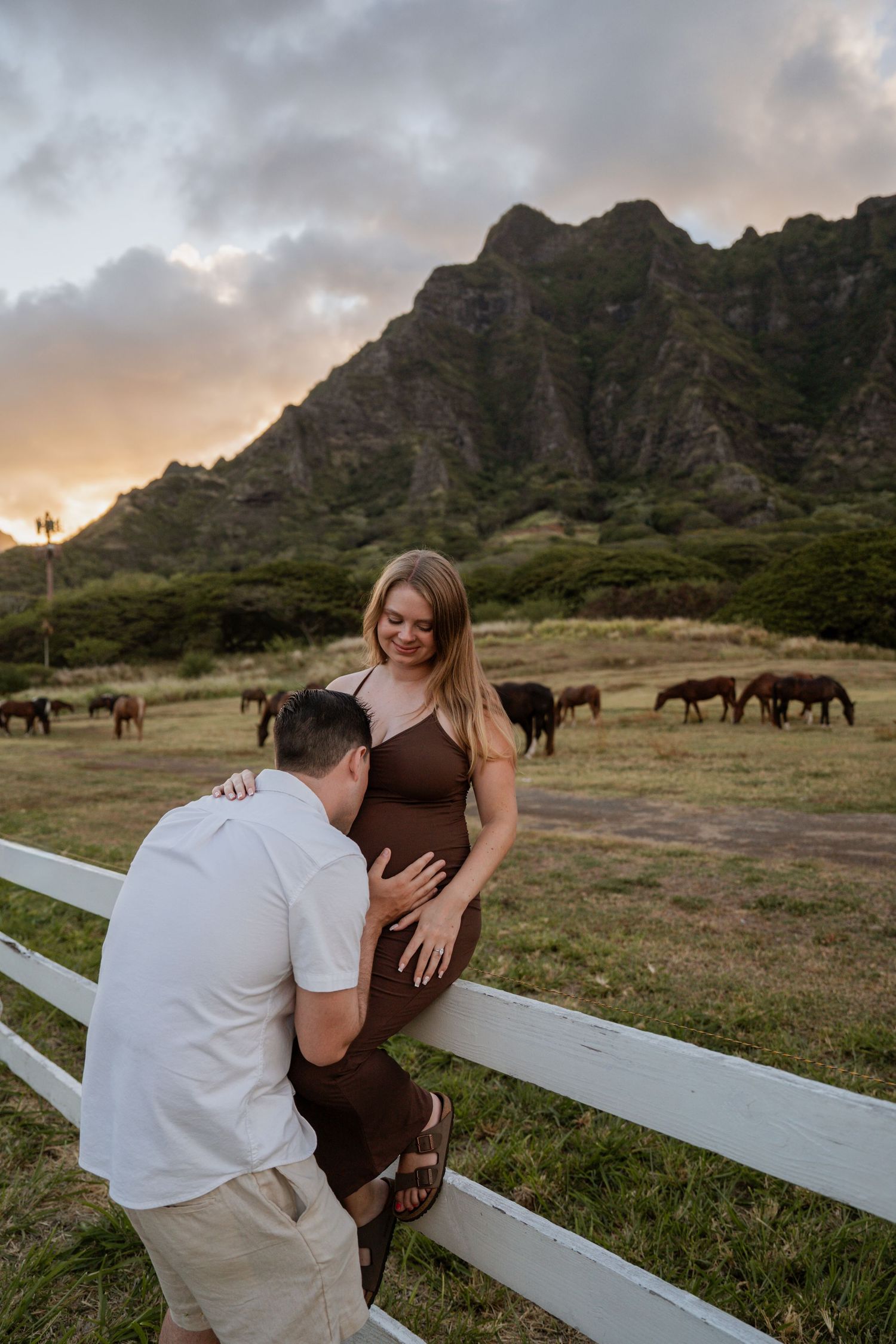 A romantic couple shares an intimate moment by a white fence at sunset with wild horses grazing in the Hawaiian mountains. Oahu Hawaii photo