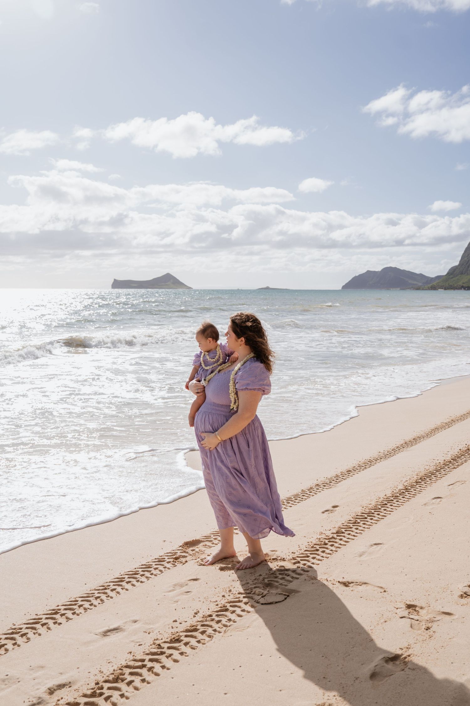 Pregnant woman in purple dress holds child on sunny Hawaiian beach with ocean waves and mountain islands in background.