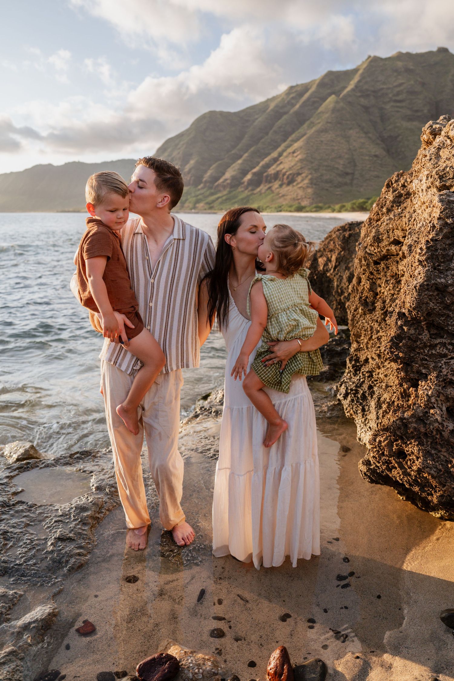 Family of four poses on rocky Hawaiian beach at sunset, parents kissing children with lush green mountains in background.